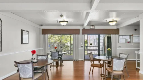 A dining room with round wooden tables, chairs with patterned backs, and large windows leading to an outdoor patio