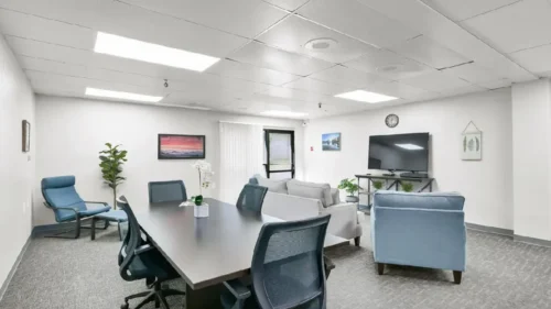 A meeting or conference room with a large dark table and rolling chairs, a light gray couch, and two light blue armchairs, with a television mounted on the wall
